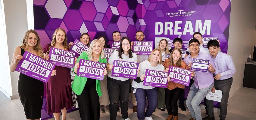 Students gather in front of a Des Moines University backdrop, proudly holding purple “I Matched in Iowa” signs.