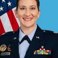 AwomaninamilitaryuniformsmilesinaformalportraitwithanAmericanflaginthebackground