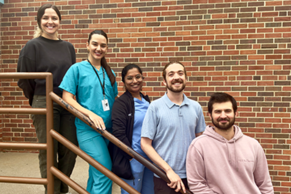 Five people stand on a staircase against a brick wall, smiling. They appear diverse and relaxed, with one in medical scrubs.