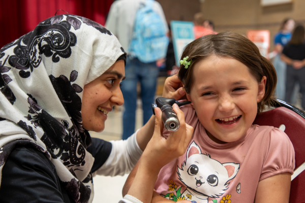 A woman wearing a patterned hijab uses an otoscope to check a smiling girl's ear in a playful setting. The girl wears a pink cat t-shirt.