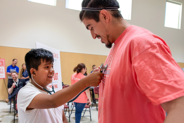 A boy at a health fair uses a stethoscope to listen to a man's heartbeat.