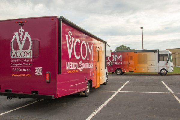 Two VCOM Mobile Medical Outreach vans in a parking lot, featuring vibrant branding with maroon and orange colors, parked under a cloudy sky.