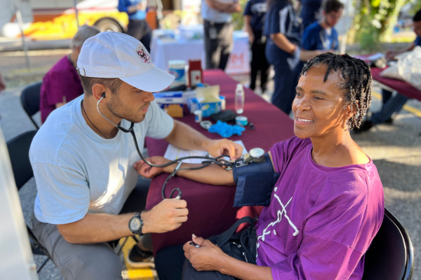 A medical student checks a smiling woman's blood pressure outdoors at a community health event.