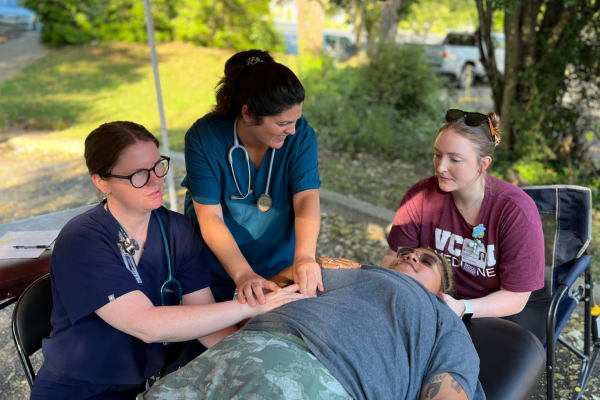 Three healthcare workers conduct osteopathic manipulative treatment on a person lying on a stretcher under a canopy outdoors. The team appears focused and caring.